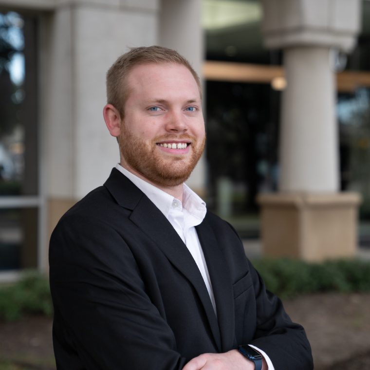 A confident man in a black suit smiles outdoors.