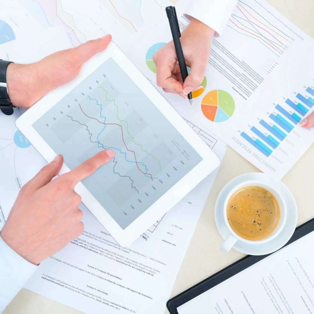 Hands analyzing financial charts on a tablet with documents and coffee nearby.